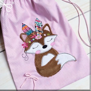 May include: A pink drawstring bag with a white polka dot pattern. The bag features an embroidered fox with brown fur, closed eyes, and a colorful feather headdress.