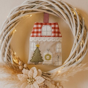May include: A white wicker wreath with a decorative Christmas house ornament. The ornament is beige with a red and white checkered roof, a green Christmas tree, and a yellow star. The wreath is decorated with dried flowers and fairy lights.