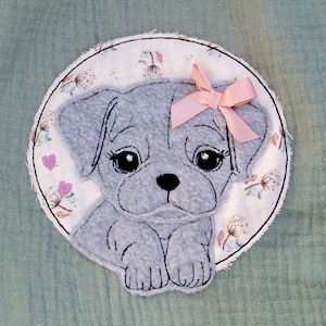 May include: A light green muslin fabric with a circular embroidered patch featuring a gray puppy with a pink bow. The puppy is surrounded by a white background with small pink hearts and white flowers.