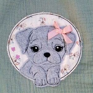 May include: A light green muslin fabric with a circular embroidered patch featuring a gray puppy with a pink bow. The puppy is surrounded by a floral design.