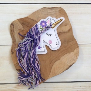 Könnte beinhalten: Weißer Filz-Einhorn-Applikation mit einer lila und silbernen Garnmähne und einem Schwanz. Das Einhorn hat ein goldenes Horn und eine Blumenkrone mit lila Blumen.