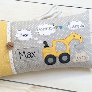 May include: A gray and yellow baby pillow with a yellow excavator, clouds with embroidered text, and the name Max. The text on the clouds reads "54cm", "02.01.2025", "13:08 Uhr", and "3370g".