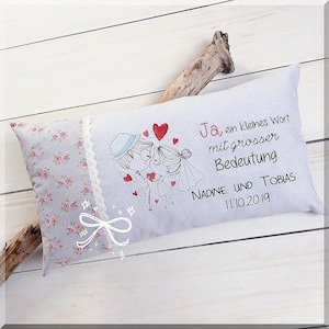 May include: A white pillow with a floral pattern on one side and a gray side with an embroidered couple, hearts, and the text "Ja, ein kleines Wort mit grosser Bedeutung NADINE UND TOBIAS 11.10.2019".