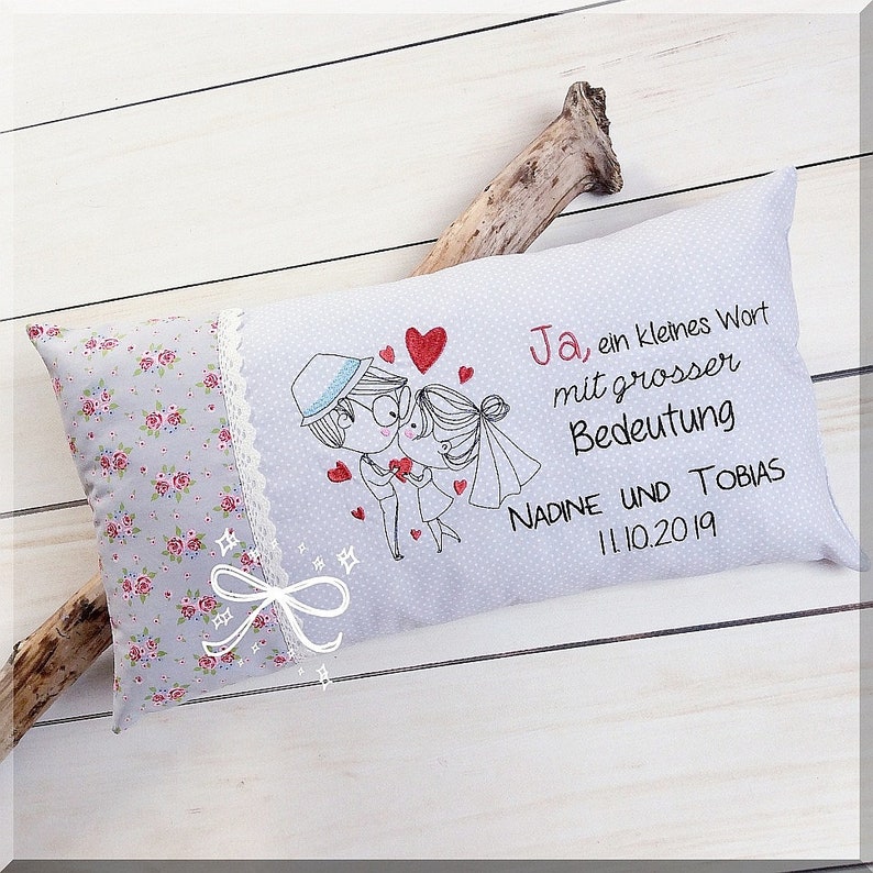 May include: A white pillow with a floral pattern on one side and a gray side with an embroidered couple, hearts, and the text "Ja, ein kleines Wort mit grosser Bedeutung NADINE UND TOBIAS 11.10.2019".