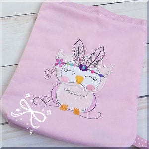 Può includere: Una borsa di tessuto rosa con un gufo ricamato che indossa una fascia di piume colorate. Il gufo ha gli occhi chiusi e un becco giallo.