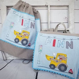 Könnte beinhalten: Zwei beige und blaue Stofftaschen mit einem blau-weiß karierten Muster. Die Taschen haben einen blauen gewellten Rand und tragen den Namen "Finn" in bunten Buchstaben. Eine Tasche hat einen gelben Spielzeug-LKW mit einer roten und weißen Ampel. Die Taschen sind perfekt für die Sachen eines Kindes.