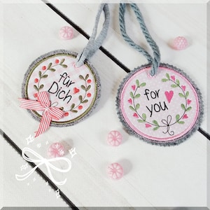 Puede incluir: Dos adornos redondos de fieltro con bordados en rosa y verde. Un adorno dice "For You" con un corazón rosa. El otro adorno dice "Für Dich" con un corazón rojo.