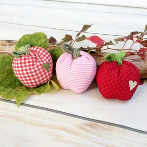 May include: Three fabric apple decorations in red gingham, pink stripes, and red polka dots. Each apple has a green stem and a small green heart appliqué. The apples are arranged on a wooden surface with leaves and branches.