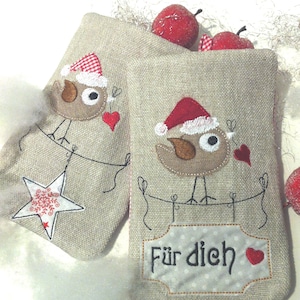 Könnte beinhalten: Zwei beigefarbene Leinensäckchen mit gestickten roten Vögeln, die Weihnachtsmützen tragen. Die Säckchen haben einen weißen Stern und ein Schild mit der Aufschrift "Für dich" mit einem roten Herzen.