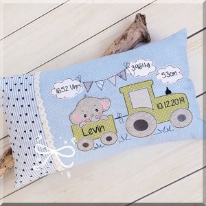 May include: A light blue pillow with a white and blue polka dot pattern. The pillow features an embroidered design of a gray elephant riding a green tractor with the text "Levin" and the date "10.12.2019".