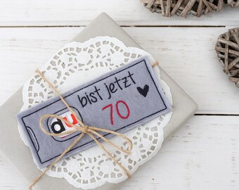 Può includere: Un regalo confezionato con un'etichetta grigia che recita "bist jetzt 70" in nero e rosso, con un piccolo cuore nero. Il regalo è legato con spago e poggia su un centrino bianco. Due decorazioni a forma di cuore intrecciate sono sullo sfondo.