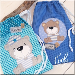 May include: Two blue drawstring bags with a bear design. One bag has a blue and white striped bear with a sailor hat and the other has a brown bear with a blue and white striped shirt. Both bags have the name "Levin" on them. One bag also has the word "Cool" on it.