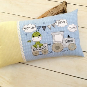 May include: A light blue pillow with a white polka dot pattern features a green dinosaur riding a grey toy tractor. The pillow has white lace trim and includes the text "14:02 Uhr", "10.12.2020", "3652g", "53cm", and "Samu".
