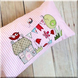 May include: A pink and white striped pillow with a house, a bird, a flower, and a butterfly applique. The pillow has a whimsical, cottagecore aesthetic.