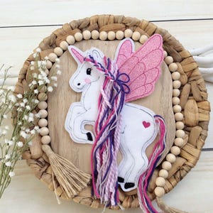 Könnte beinhalten: Weißes Filz-Einhorn mit rosa Glitzerflügeln und einer rosa, lila und weißen Garnmähne und -schwanz. Das Einhorn befindet sich auf einem Holzgrund mit einem braunen Quasten.