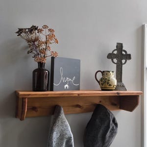 Puede incluir: Una estantería de madera con un jarrón marrón oscuro con flores secas, un letrero gris con la palabra "home", una cruz decorativa y una pequeña jarra de cerámica. Debajo, un suéter gris y una gorra gris cuelgan de ganchos.