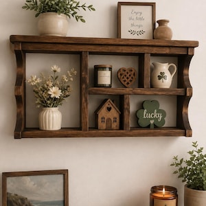 May include: A dark brown wooden wall shelf with multiple compartments holding a small potted plant, a framed sign reading Enjoy the little things, a candle, a wooden heart, a small pitcher, a wooden house, and a green shamrock sign labeled lucky.