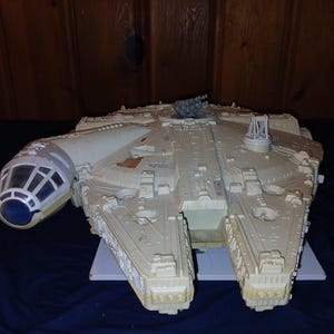 May include: A detailed model of the Millennium Falcon starship, featuring a light beige color scheme with gray and blue accents. The model includes intricate detailing, a cockpit, and various turrets, showcasing its iconic design from the Star Wars franchise.