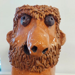 May include: A ceramic sculpture of a face with a large nose, bushy eyebrows, and a beard. The sculpture is painted in shades of brown and has blue eyes.