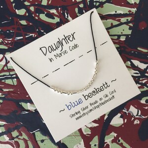 Puede incluir: Una tarjeta blanca con texto negro que dice "Daughter in Morse Code" y un cordón negro con cuentas de plata que deletrean la palabra "daughter" en código Morse. La tarjeta también incluye el texto "~ blue beckett ~ Sterling Silver Beads on Silk Cord www.etsy.com/shop/bluebeckett".