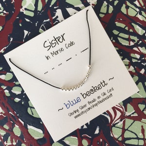 Puede incluir: Una tarjeta blanca con texto negro que dice "Hermana en código Morse" y un collar de cuentas de plata sobre un cordón negro. La tarjeta también incluye el texto "~ blue beckett ~ Cuentas de plata de ley en cordón de seda www.etsy.com/shop/bluebeckett"