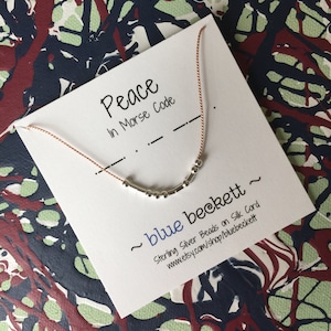 Puede incluir: Un collar de plata con cuentas que deletrean la palabra "paz" en código Morse en una tarjeta blanca con el texto "Peace in Morse Code" y "~ blue beckett ~ Sterling Silver Beads on Silk Cord www.etsy.com/shop/bluebeckett".