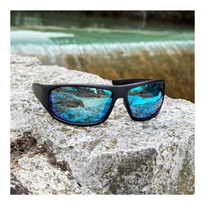 Könnte beinhalten: Schwarze Sonnenbrille mit blau verspiegelten Gläsern liegt auf einem grauen Felsen. Die Gläser spiegeln eine lebendige türkise und blaue Landschaft wider. Die Sonnenbrille hat einen dicken, rechteckigen Rahmen. Ideal für Outdoor-Aktivitäten.