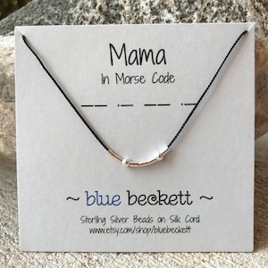 Puede incluir: Un collar de cordón negro con cuentas de plata que deletrean "Mama" en código Morse. El collar se muestra en una tarjeta blanca con el texto "Mama in Morse Code" y "~ blue beckett ~ Sterling Silver Beads on Silk Cord www.etsy.com/shop/bluebeckett".