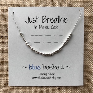 May include: A silver necklace with a beaded chain and a message that reads "Just Breathe" in Morse code. The necklace is displayed on a white card with the text "blue beckett" and "Sterling Silver" printed on it.