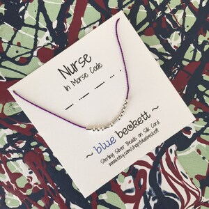 Puede incluir: Un collar de cordón morado con cuentas de plata que deletrean "Nurse" en código Morse. El collar se muestra en una tarjeta blanca con el texto "Nurse in Morse Code" y "~ blue beckett ~ Sterling Silver Beads on Silk Cord www.etsy.com/shop/bluebeckett".