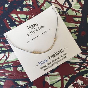 Puede incluir: Una tarjeta blanca con la palabra "Hope" escrita en tinta negra, seguida de "In Morse Code" y una serie de guiones y puntos. La tarjeta también presenta un collar de cuentas de plata y oro con el texto "~blue beckett~ Sterling Silver Beads on Silk Cord www.etsy.com/shop/bluebeckett~" escrito en tinta negra.