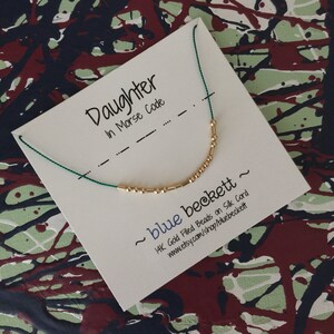 Puede incluir: Una tarjeta blanca con el texto "Daughter in Morse Code" y un collar de cuentas doradas en un cordón verde. La tarjeta también incluye el texto "~ blue beckett ~ 14K Gold Filled Beads on Silk Cord www.etsy.com/shop/bluebeckett".