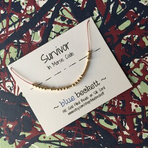Puede incluir: Una tarjeta blanca con el texto "Survivor in Morse Code" y "~ blue beckett ~ 14K Gold Filled Beads on Silk Cord www.etsy.com/shop/bluebeckett" impreso en ella. Una pulsera de cuentas doradas se muestra en la tarjeta.
