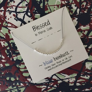 May include: A silver beaded necklace on a silk cord, spelling "Blessed" in Morse code. The necklace is displayed on a white card with the text "Blessed In Morse Code" and "blue beckett". The background has a colorful abstract design.