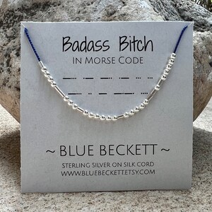 Puede incluir: Un brazalete de cuentas de plata con un cordón azul, que deletrea "Badass Bitch" en código Morse en una tarjeta blanca con el texto "Badass Bitch in Morse Code", "~ BLUE BECKETT ~", "STERLING SILVER ON SILK CORD" y "WWW.BLUEBECKETT.ETSY.COM".