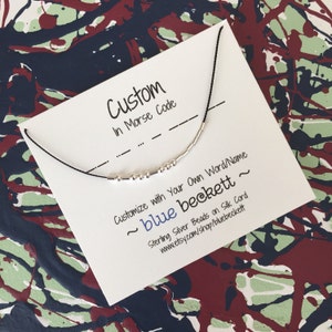 May include: A white card with black text that reads "Custom in Morse Code" and "Customize with your own word/name ~ blue beckett ~ Sterling Silver Beads on Silk Cord www.etsy.com/shop/bluebeckett". A black cord with silver beads is strung through holes in the card.