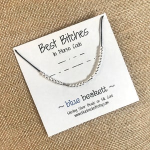 Op de afbeelding: Een witte kaart met zwarte tekst die "Best Bitches in Morse Code" zegt en een zilveren kralenarmband op een zwart koord. De kaart bevat ook de tekst "~ blue beckett ~ Sterling Silver Beads on Silk Cord www.bluebeckett.etsy.com".