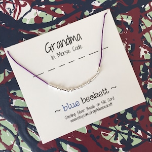 Puede incluir: Una tarjeta blanca con la palabra "Grandma" escrita en tinta negra. La tarjeta también incluye la frase "In Morse Code" y una serie de guiones y puntos. Un brazalete plateado con pequeñas cuentas se muestra en la tarjeta. La tarjeta también incluye el texto "~ blue beckett ~ Sterling Silver Beads on Silk Cord www.etsy.com/shop/bluebeckett"