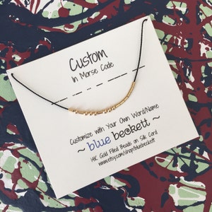 Può includere: Una collana con cordoncino nero e perline dorate che compongono la parola "blue beckett" in codice Morse. La collana è esposta su un cartoncino bianco con il testo "Custom in Morse Code" e "Customize with your own word/name ~ blue beckett ~ 14K Gold Filled Beads on Silk Cord www.etsy.com/shop/bluebeckett".