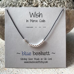 Puede incluir: Un collar de cordón negro con cuentas de plata que deletrean la palabra "wish" en código Morse. La tarjeta dice "Wish in Morse Code" y "~blue beckett~ Sterling Silver Beads on Silk Cord www.bluebeckett.etsy.com".
