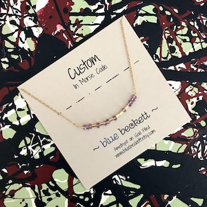 May include: A close-up of a card with the text "Custom in Morse Code" and a necklace with amethyst beads and gold spacers. The card also includes the text "~blue beckett~ Amethyst on Gold Filled www.bluebeckett.etsy.com"