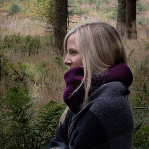 May include: A woman wearing a purple neck warmer and a gray plaid shawl stands in a forest setting. She is looking off to the side, and her blonde hair is flowing in the wind.