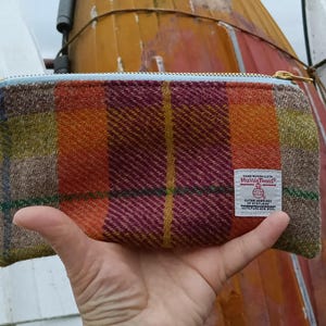 Op de afbeelding: Een klein ritsvakje gemaakt van een kleurrijke geruite stof. Het vakje heeft een bruin, oranje, geel en rood geruit patroon. Het vakje heeft een witte rits en een klein wit label met de tekst "Made in Scotland" erop.