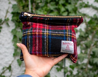 Könnte beinhalten: Eine schwarze und rote karierte Reißverschlusstasche mit einem goldenen Reißverschluss. Die Tasche hat ein weißes Etikett mit dem Text "Harris Tweed" und einem Logo, das eine Wollkugel mit einem Kreuz darin darstellt.