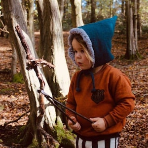 以下が含まれることがあります： 灰色のフリースの裏地が付いたターコイズブルーのフード付き帽子、パッチが付いたさび色のスウェットシャツ、白黒のストライプのパンツを着用した子供。子供は森の中で2本の棒を持っています。