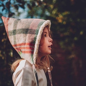 以下が含まれることがあります： ピンク、グリーン、オレンジのチェック柄のフード付き帽子をかぶった少女。帽子は白いフリースの裏地が付いており、下部に紐が付いています。