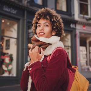 May include: A woman with curly brown hair is wearing a red corduroy jacket with a brown and white scarf. She is standing in front of a storefront with a sign that says "Walker Slater".