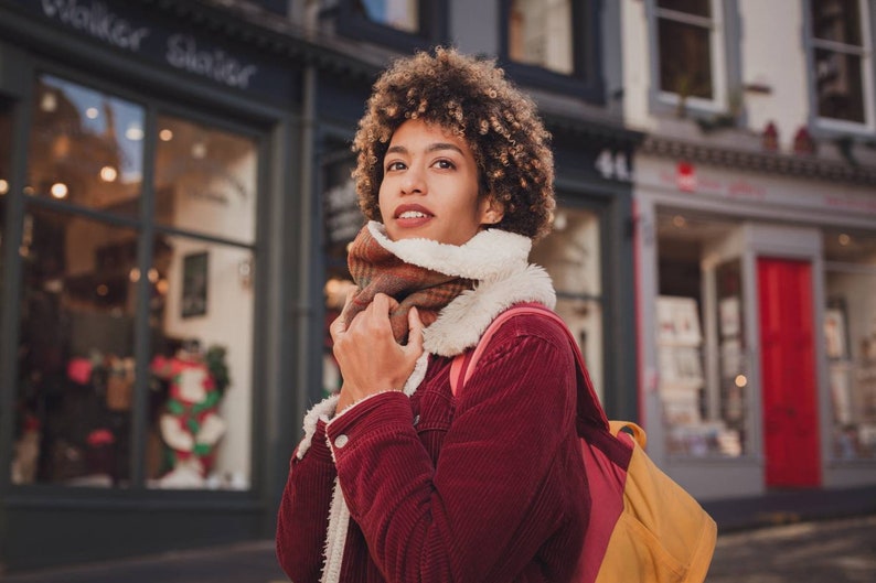 May include: A woman with curly brown hair is wearing a red corduroy jacket with a brown and white scarf. She is standing in front of a storefront with a sign that says "Walker Slater".