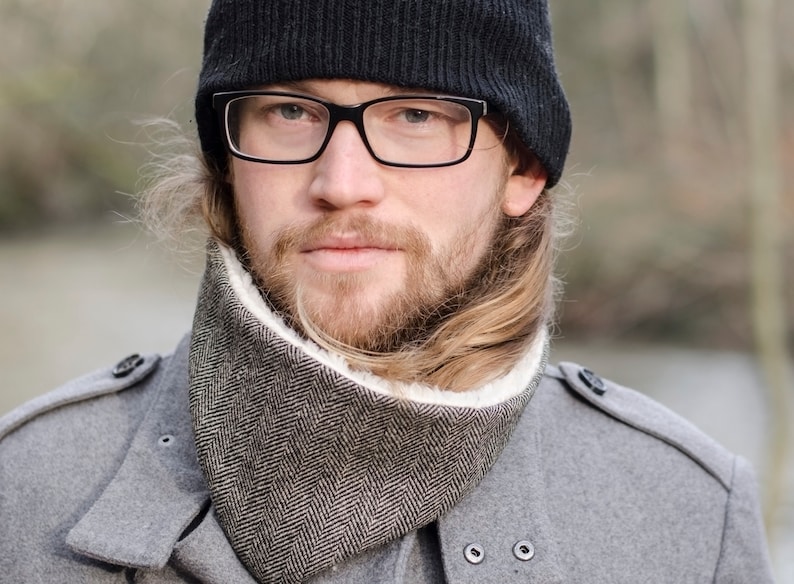 May include: A man wearing a black knit beanie, black framed glasses, a gray coat, and a brown and white patterned neck warmer.