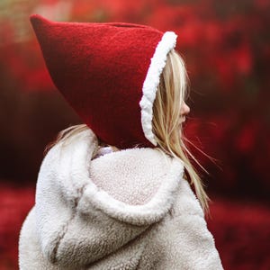 Op de afbeelding: Een jong persoon draagt ​​een rode capuchon met een witte rand en een wit pluche jack. De achtergrond is wazig rood.